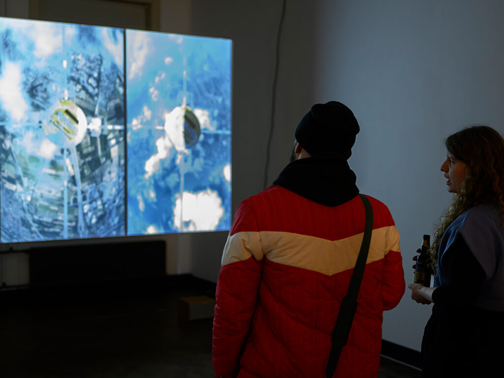 Image of exhibition work os visual media depicting a satellite view and two visitors viewing it.