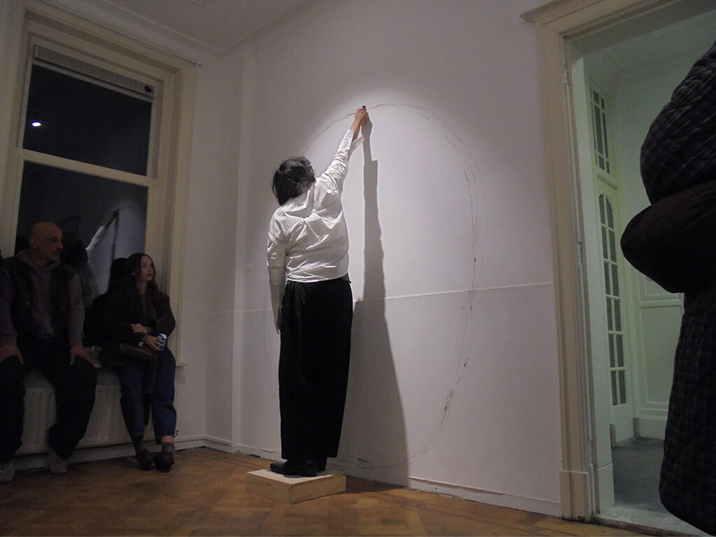 Minimalist room with a large, hand-drawn circle on a white wall made by Tom Marioni as a performative session in front of an audience.