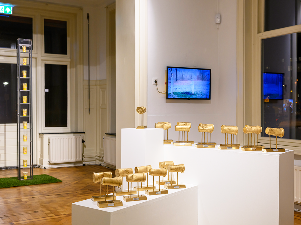 Image of the exhibition view depicting an installation with golden pudding cups and a TV in the background.