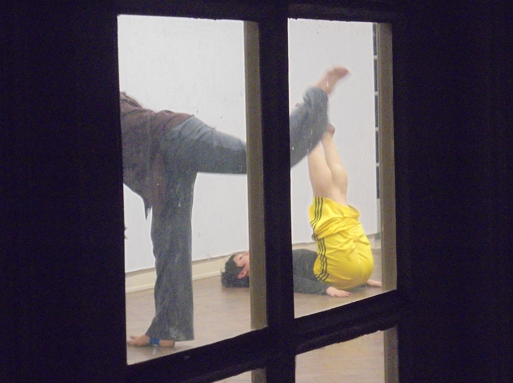 Performers improvising movements on the floor in an empty room with white walls. This image is taken outside the room.