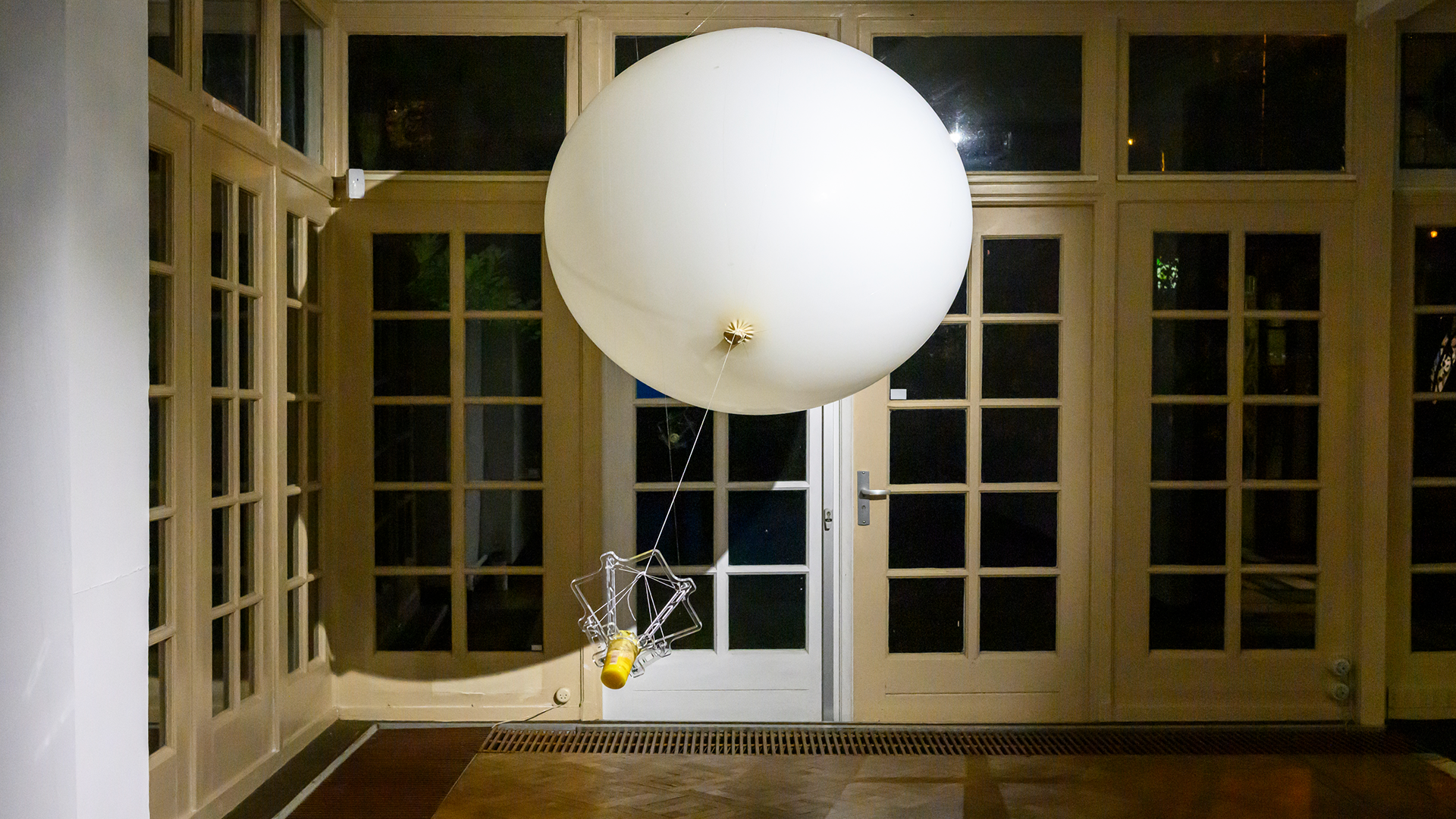 Image of the artwork or installation with big white balloon and pudding attached to it with the glass doors of the house in the background.