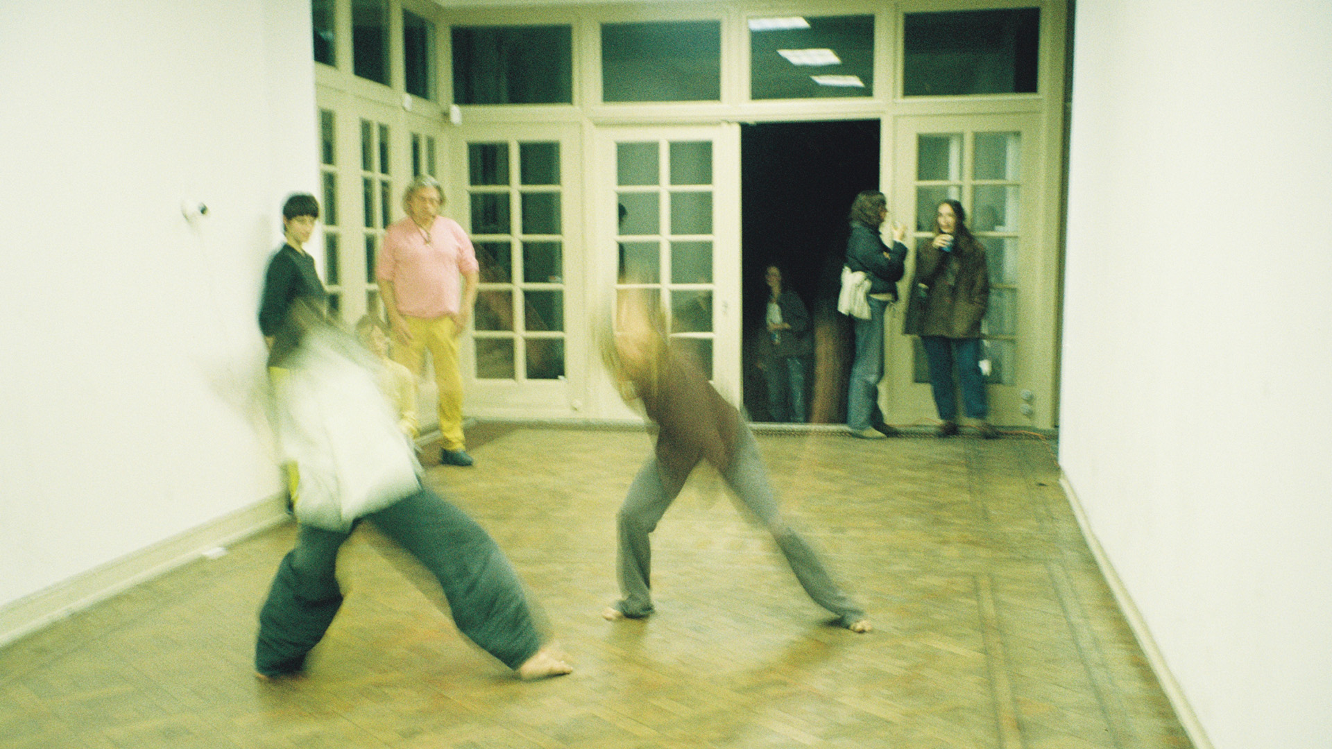 Performers improvising movements on the floor in an empty room with white walls.