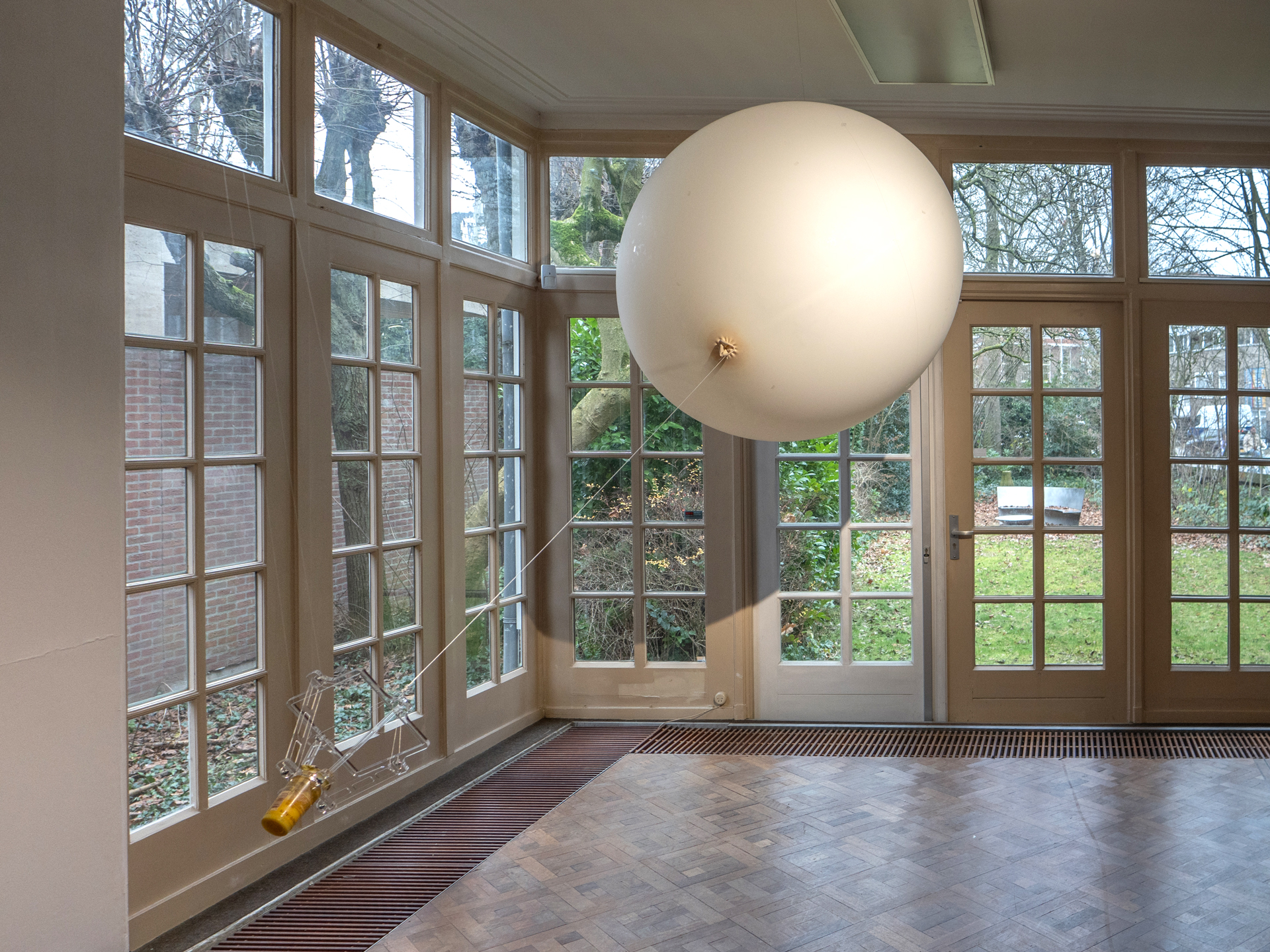 A bright room with large, grid-patterned windows showcases a floating white sphere tethered to the floor. Beyond the windows, a lush garden is visible.