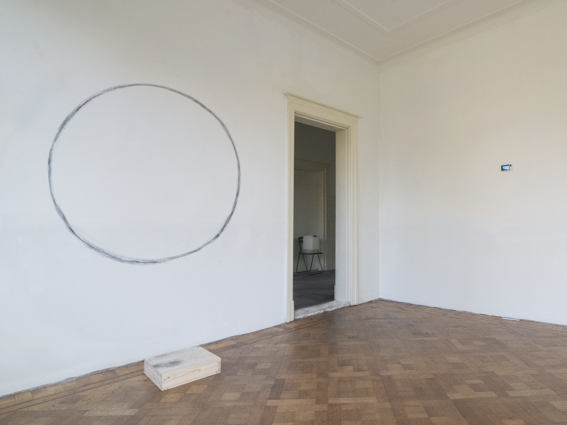 Minimalist room with a large, hand-drawn circle on a white wall, parquet flooring, a small wooden block in the corner, and an open door to an adjacent room.