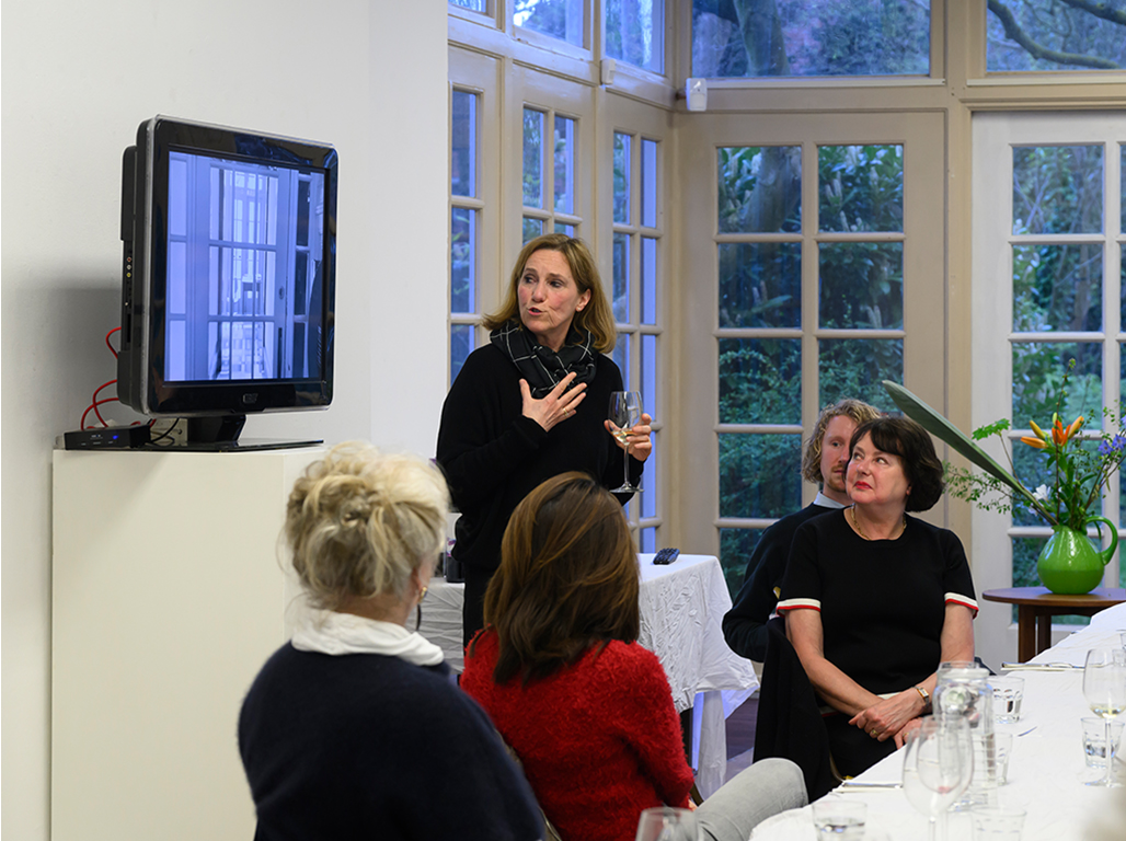 Speaker standing presenting in front of an audience seated around a table.