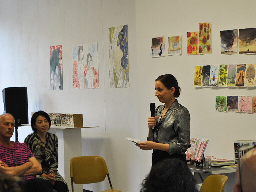 A speaker giving a lecture. Behind some paintings hanging on the wall.