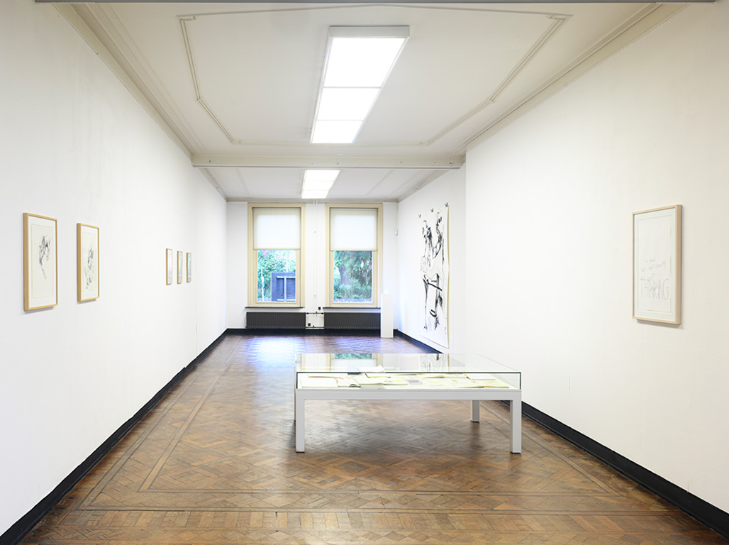 A wide white space with frames hanging from it walls and a table displaying art pieces in the centre right.