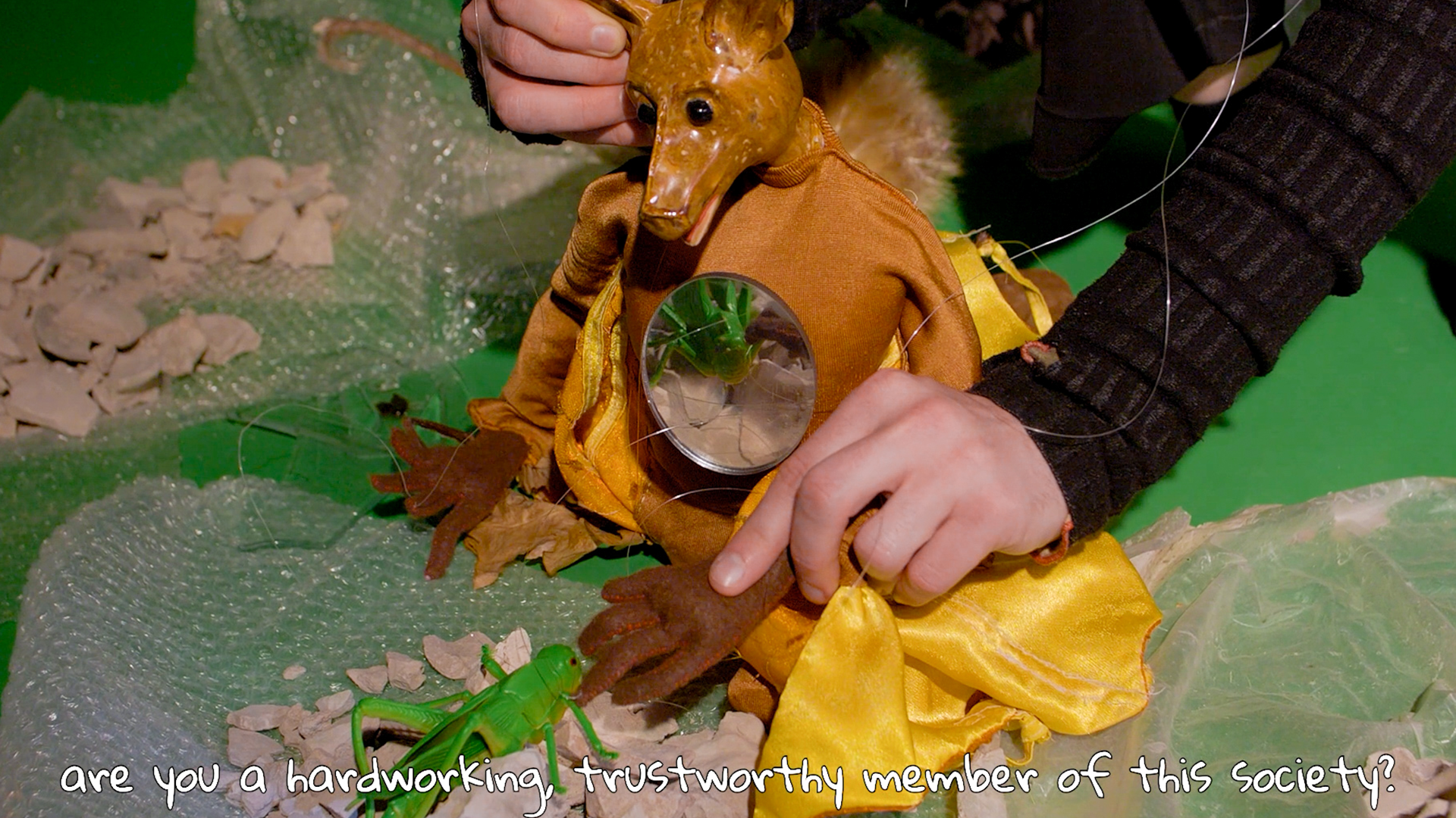 A green-and-orange composition depicting a puppet controlled by two hands, accompanied by a displayed statement