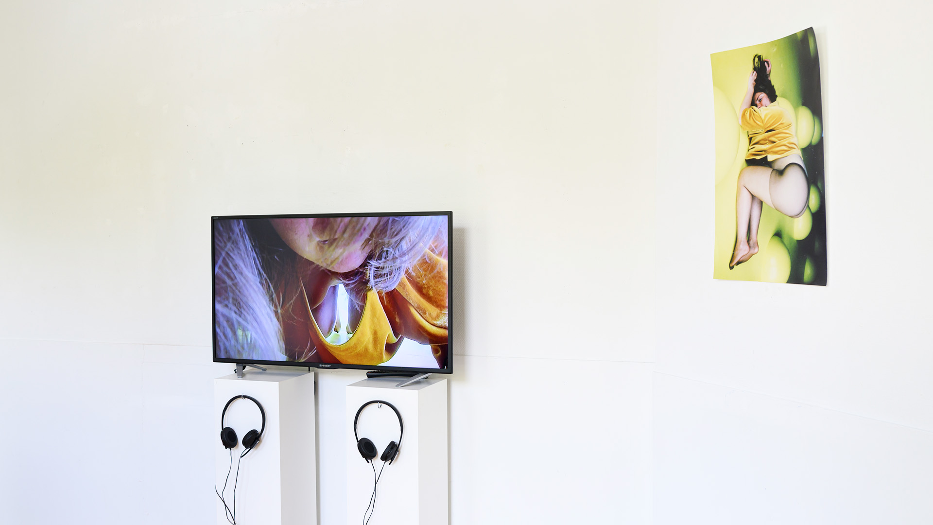 In a white room, a screen shows vibrant visuals with headphones nearby, next to a yellow photograph..