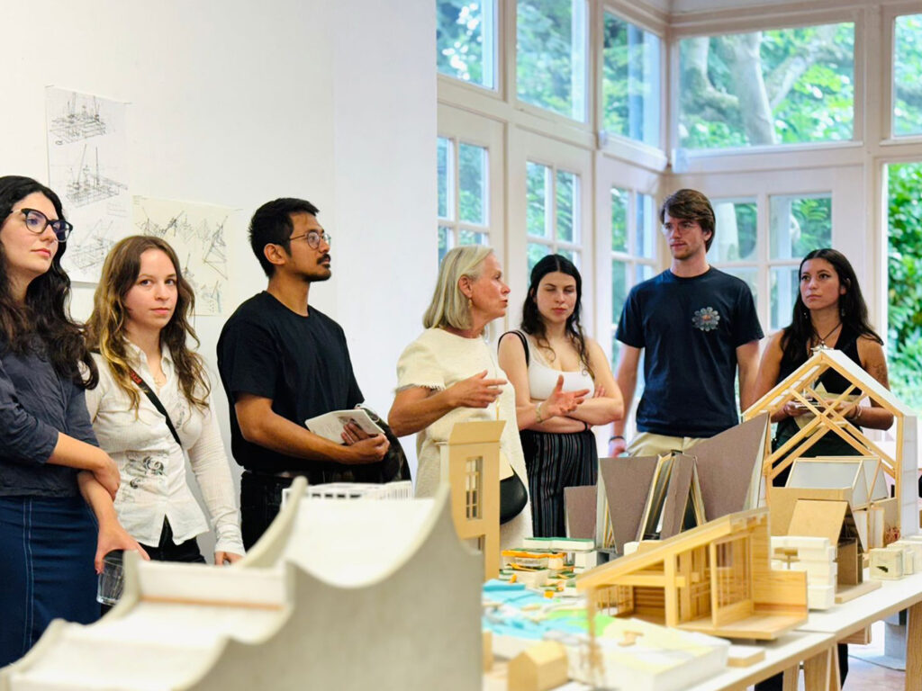Architecture maquette displayed on a table while group of students gathered around listening to a presentation.