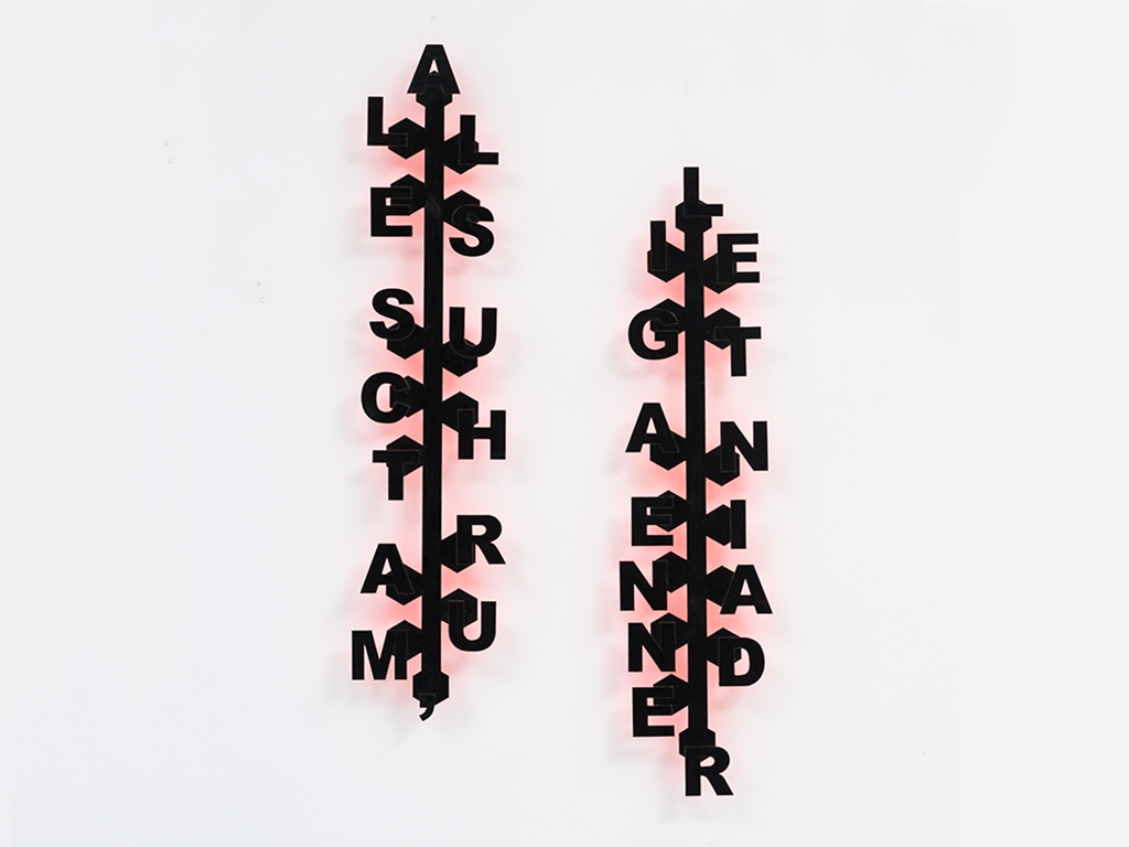 Two vertical, black typographic wall sculptures mounted on a white wall. Each piece has a central spine that connects the letters.