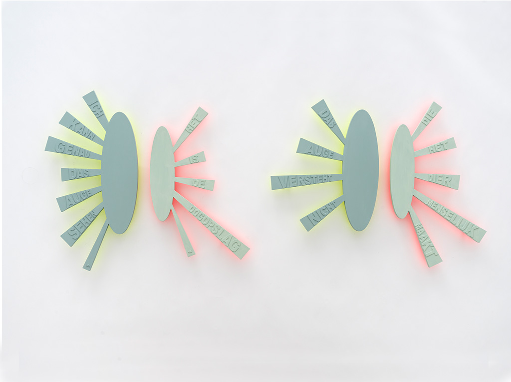 Two horizontal, green and pink typographic wall sculptures mounted on a white wall. Each piece is a composition of two round pieces.