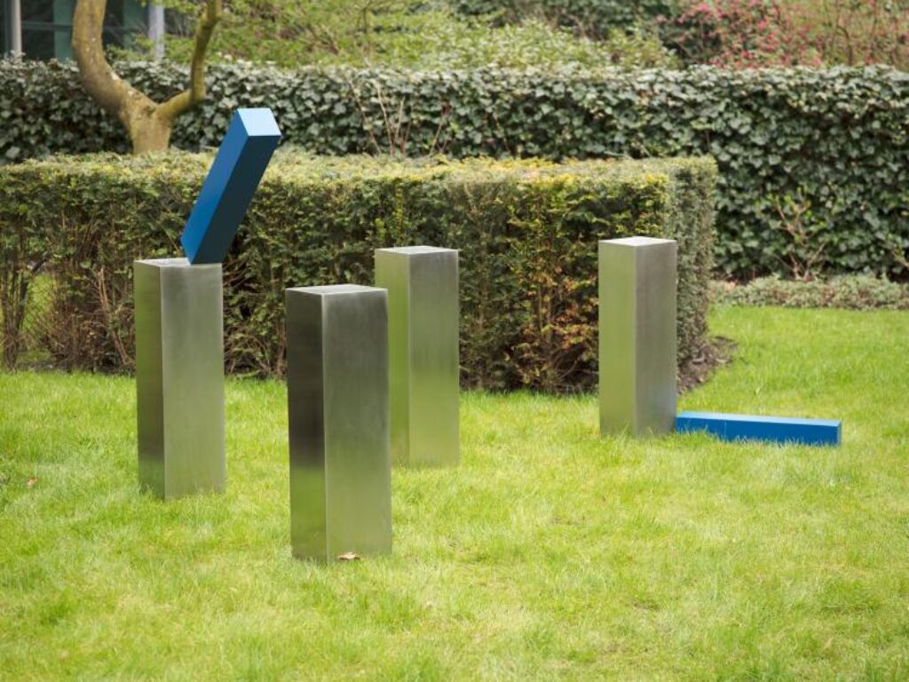 Outdoor sculpture consisting of four vertical steel poles placed in grass. Two blue steel elements are attached to the poles: one is fixed high on a pole in a tilted position, and another is attached lower to a different pole, also angled rather than straight. The structure is minimal and open, with the elements extending outward from the poles rather than forming straight connections between them.