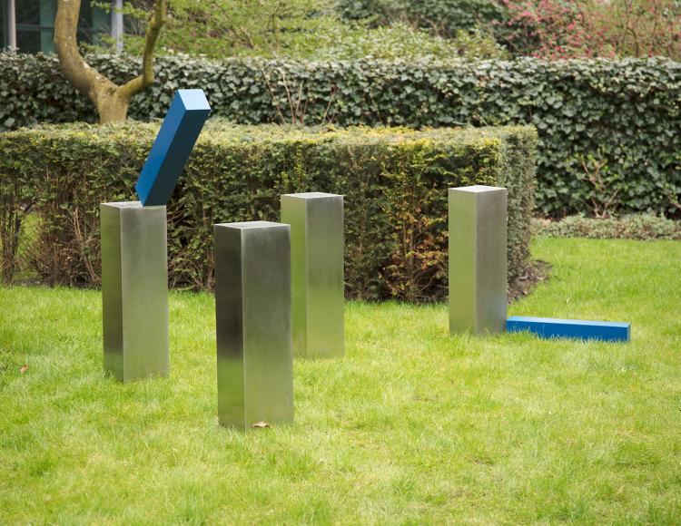 Outdoor sculpture consisting of four vertical steel poles placed in grass. Two blue steel elements are attached to the poles: one is fixed high on a pole in a tilted position, and another is attached lower to a different pole, also angled rather than straight. The structure is minimal and open, with the elements extending outward from the poles rather than forming straight connections between them.