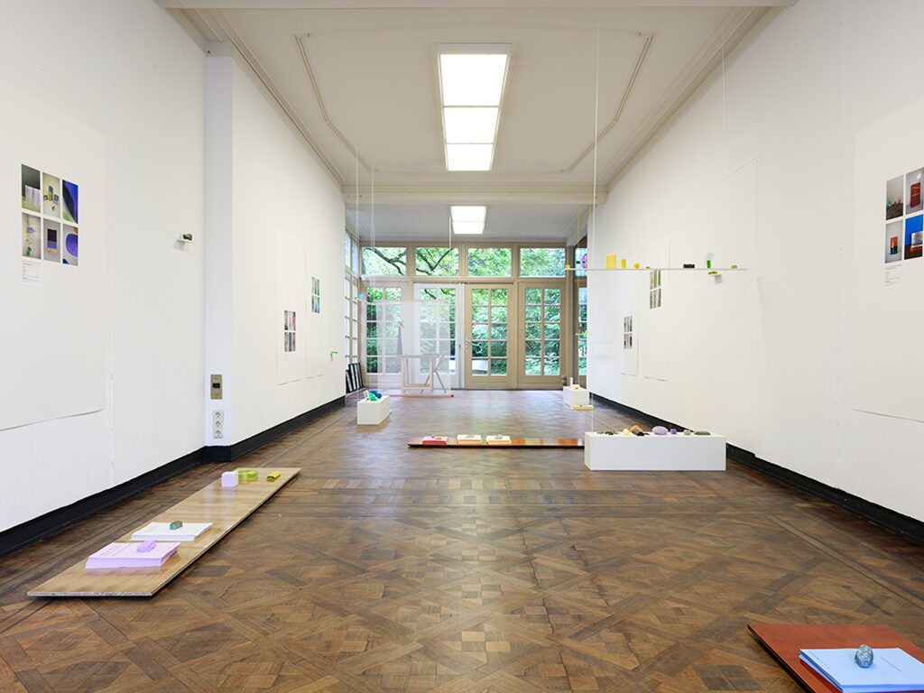 A wide room with brown floor and white walls. In the room small colourful objects scattered around.