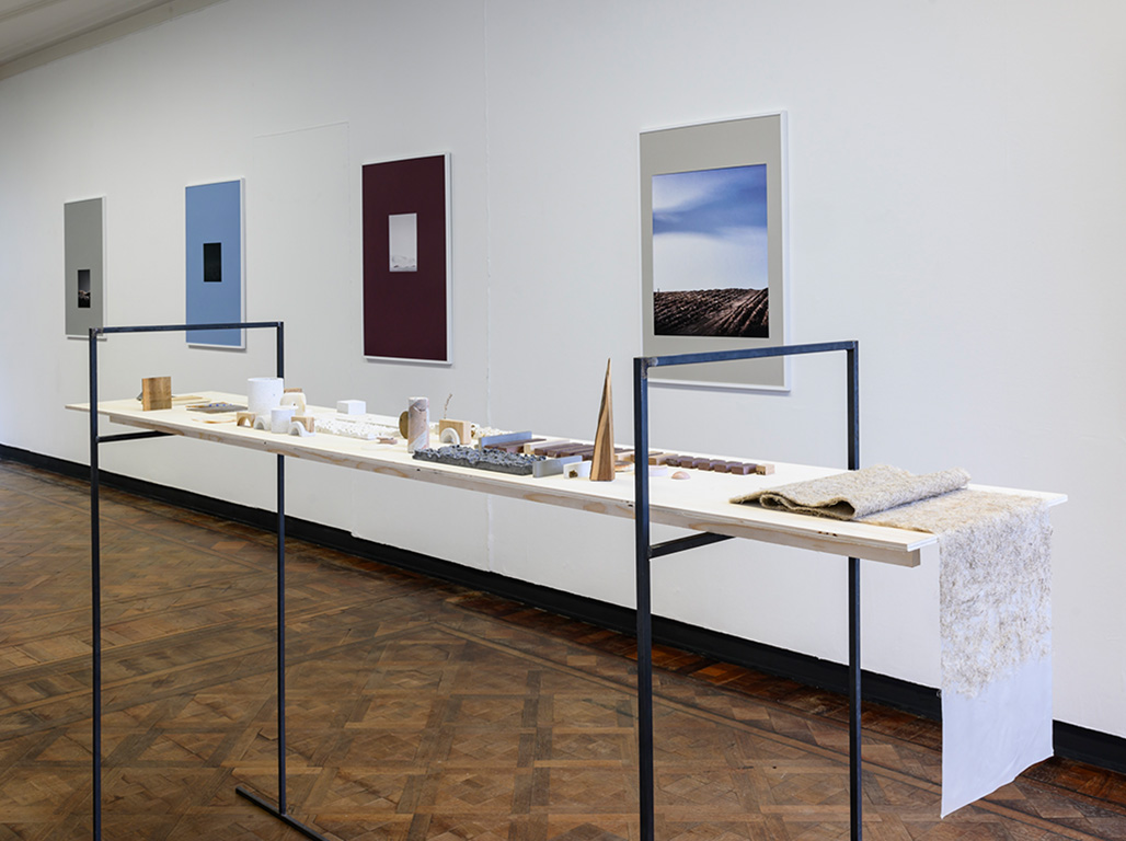A space with some blue, burgundy and grey frames hanging on the walls. In front a table made of wood and metal displaying some moquettes and sketches.