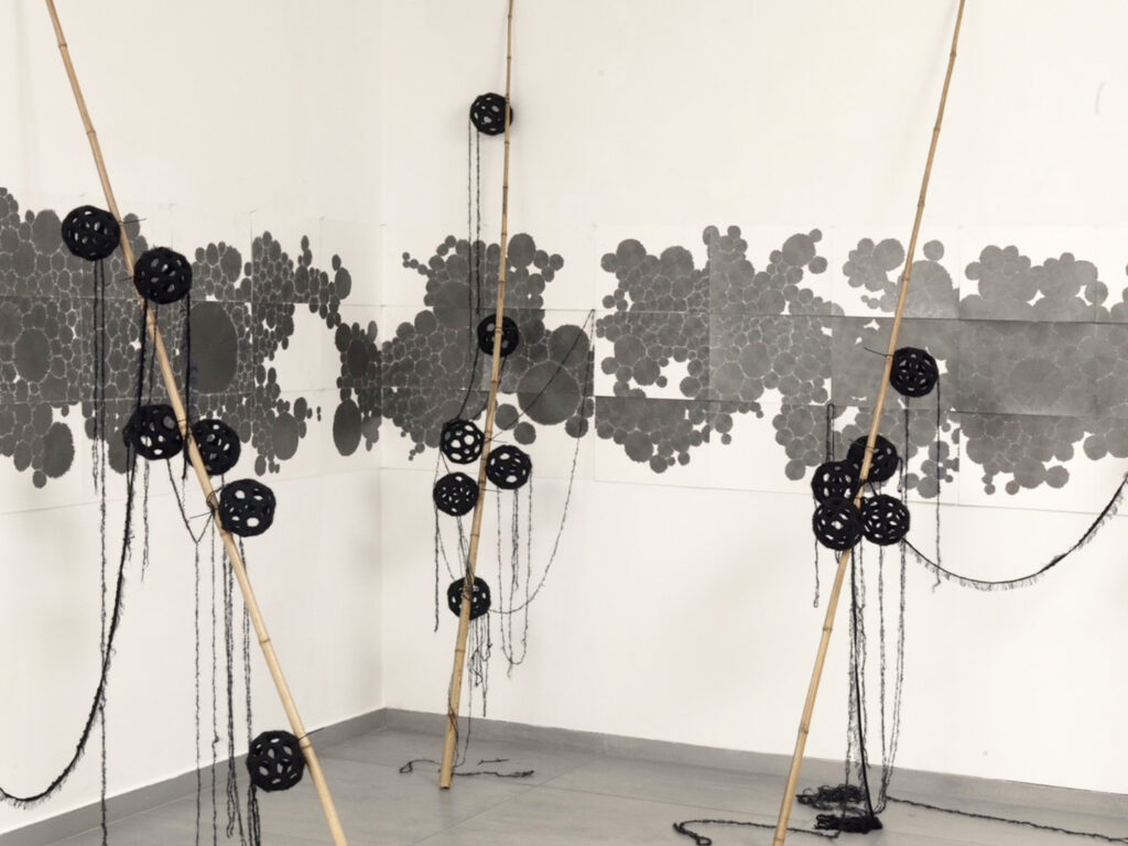 Artistic intervention on a white wall, featuring a grey, map-like composition painted across the surface, complemented by wooden sticks tipped with black spheres.