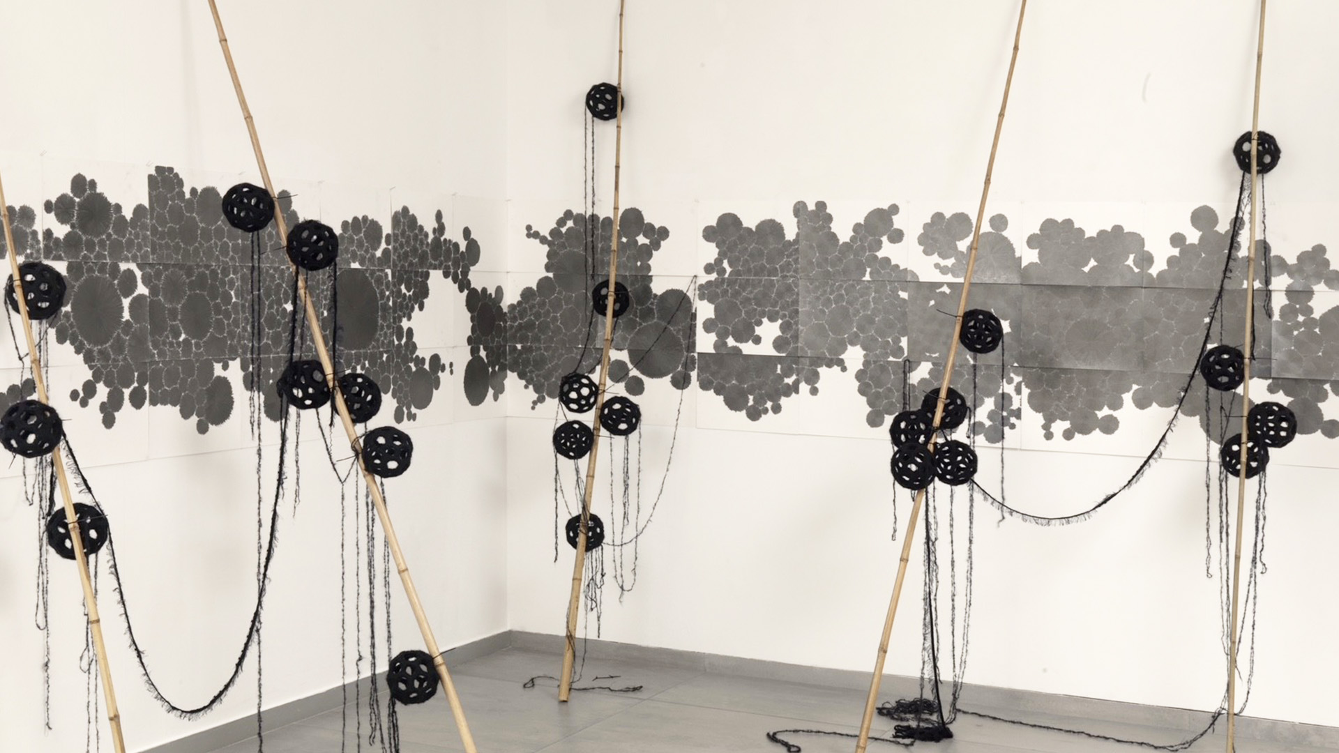 Artistic intervention on a white wall, featuring a grey, map-like composition painted across the surface, complemented by wooden sticks tipped with black spheres.