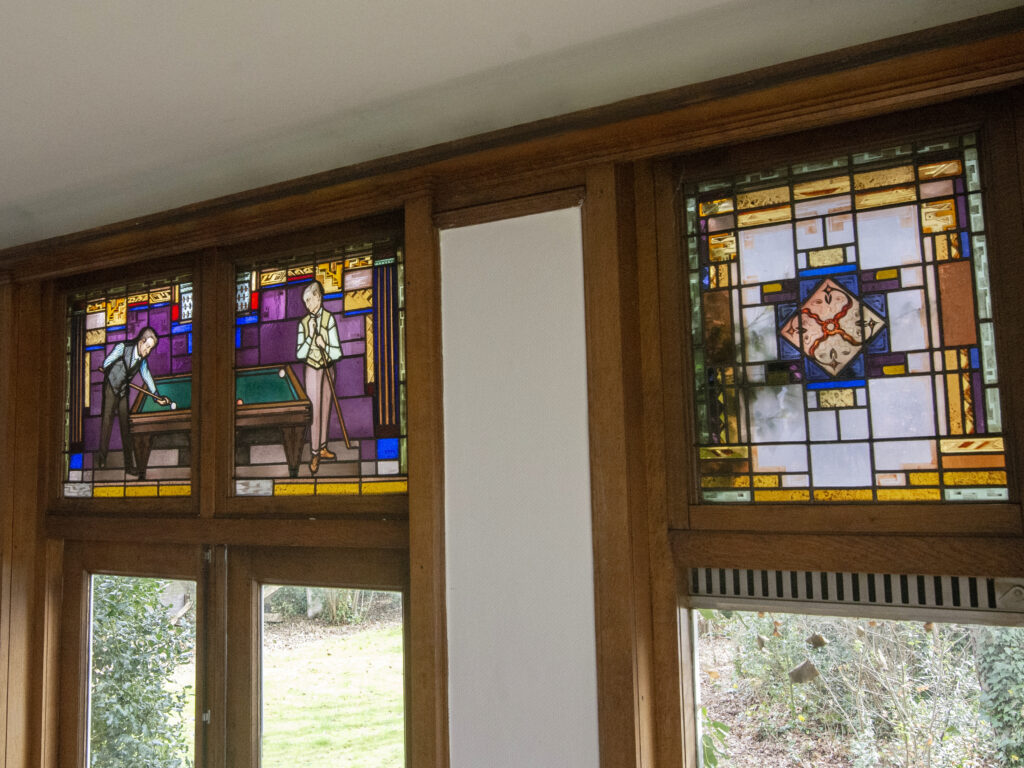 Stained-glass window in the billiard room showing two men playing billiards. One figure represents Henri van Abbe, and the other is likely Anton Philips. The scene is composed in colored glass with purple, green, pink, brown, and blue tones. The figures are positioned around a billiard table in a stylized, early 20th-century design, set within the architectural frame of the window.